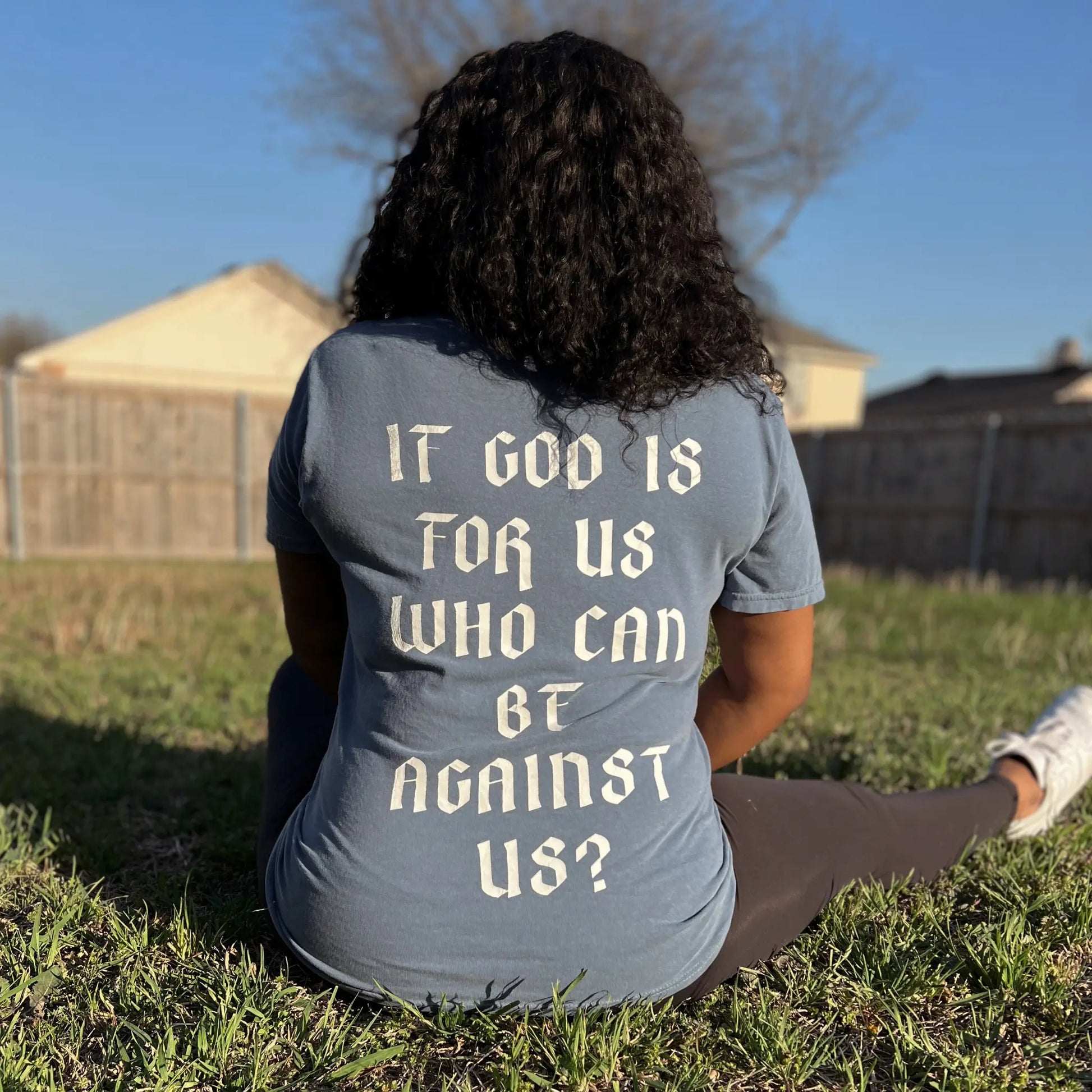Romans 8:31 T-shirt is in the color blue jean, with a crew neck style. There is a crown logo on the left side of the chest. The Romans 8:31 T-shirt is comfortable and fitted. Size up for an oversized look. On a Neutral Colored background. The back reads “If God is for us who can be against us”, in bold white letters. Photos is taken outside in the sunslight. Christian Clothing brand crown of favor