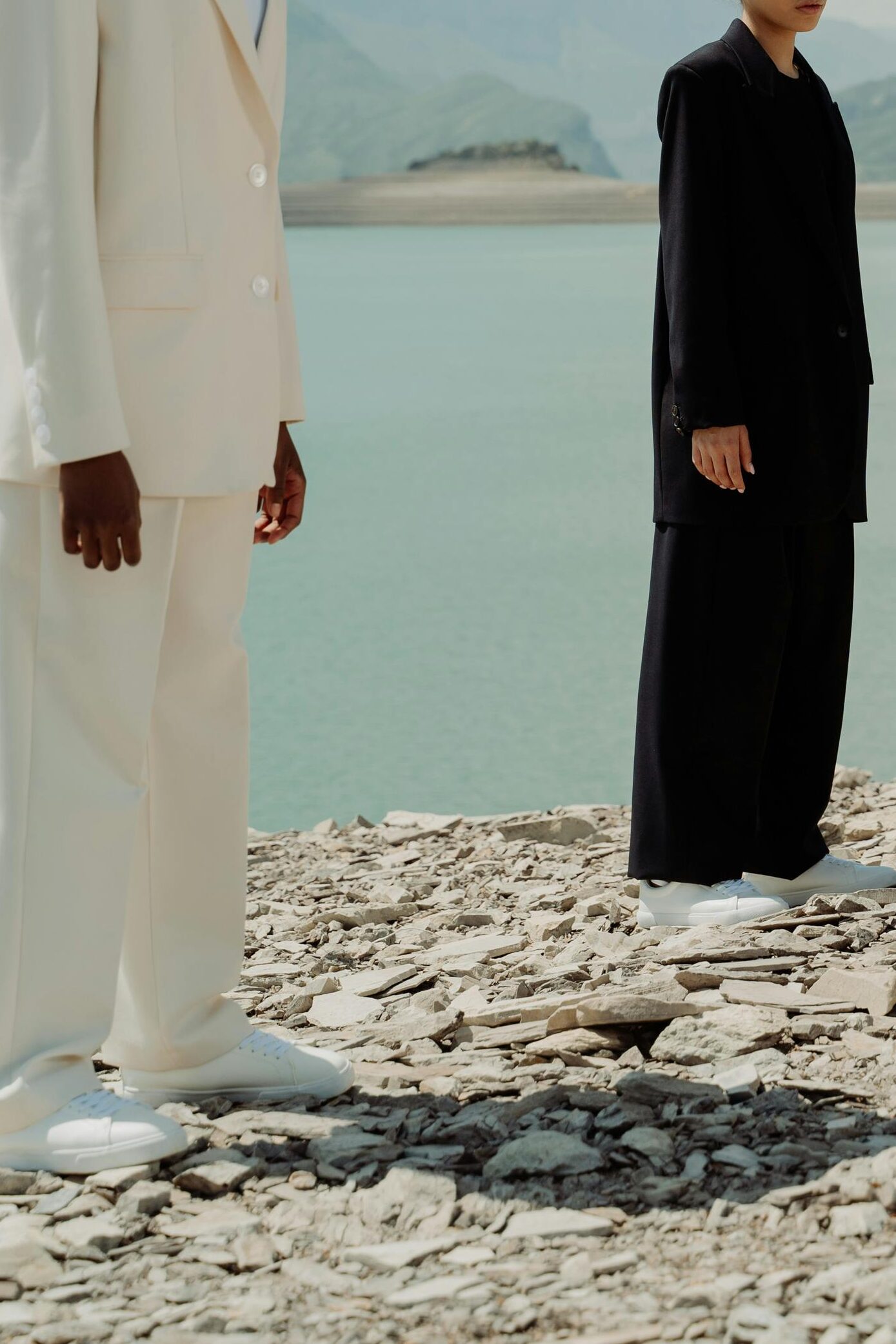 Three women in vibrant blazers stand on a rocky shore by a body of water, showcasing diversity and fashion.